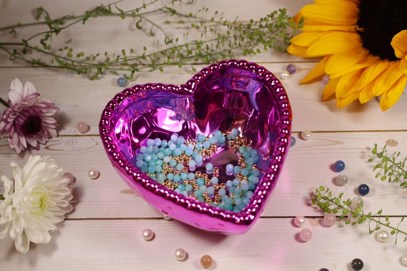 Colorful gemstone beads in a pink heart bowl surrounded by flowers and crystals